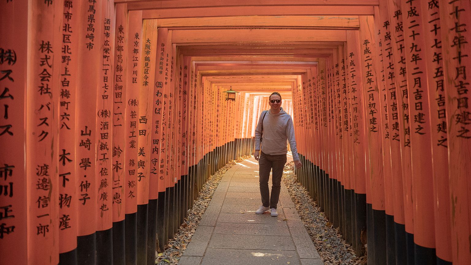 A person walks through a corridor of red torii gates with Japanese inscriptions, surrounded by sunlight and shadows.