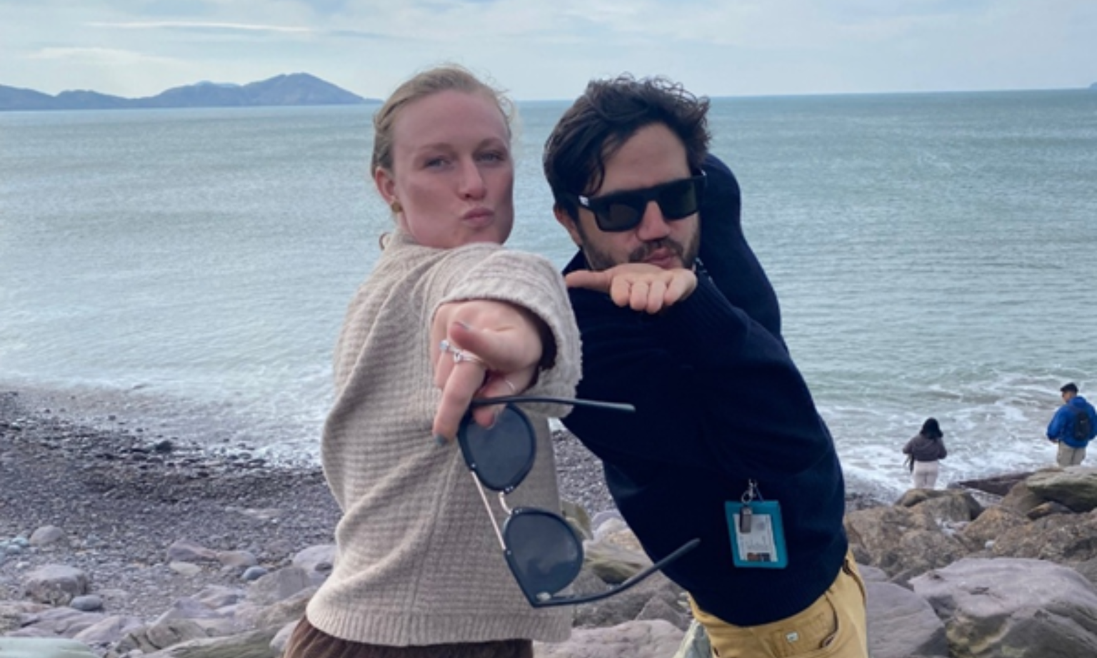 Two people striking playful poses by a rocky beach, with the ocean and distant hills in the background.