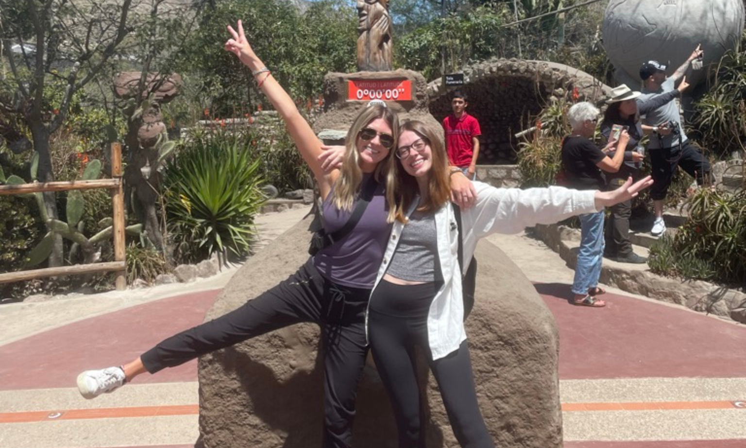 Two women smiling and posing with arms raised on a stone platform outdoors, surrounded by plants and people in the background.