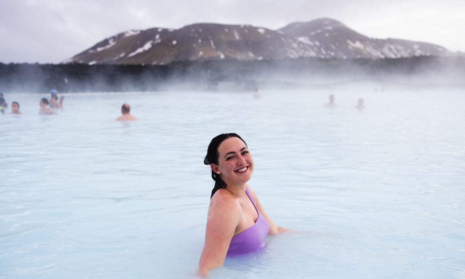 Person in a purple swimsuit smiles while standing in a steaming blue lagoon, with fog and mountainous landscape in the background.
