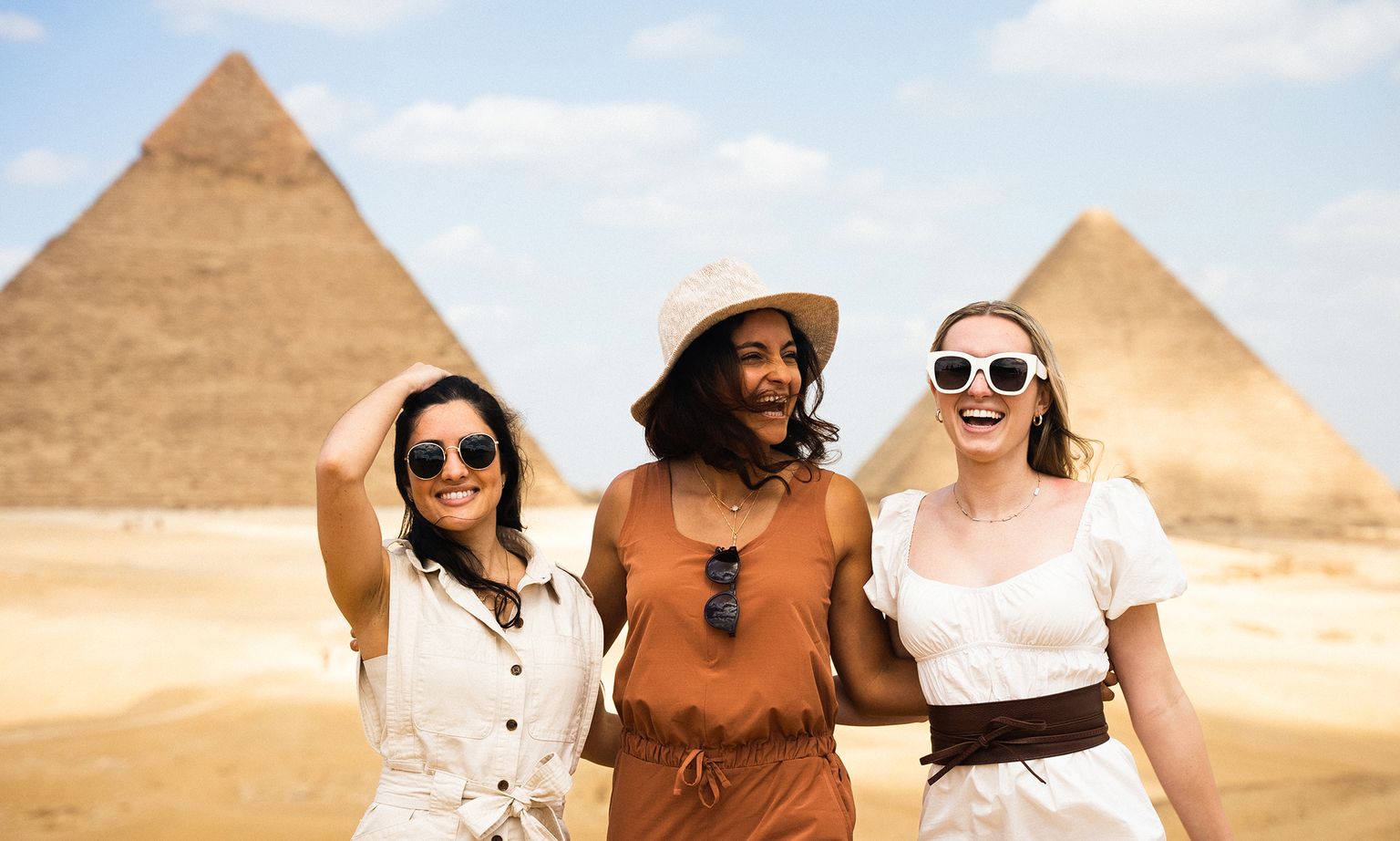 Three women smiling in front of the pyramids in Egypt, on a sunny day. They're wearing sunglasses and summer clothing.