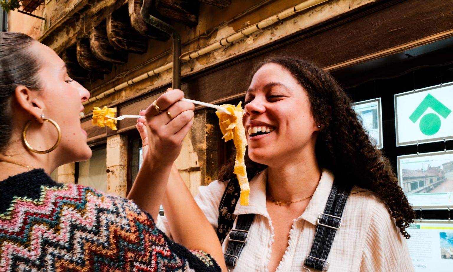 Two women laughing and feeding each other pasta with chopsticks outdoors, standing in front of a rustic building.