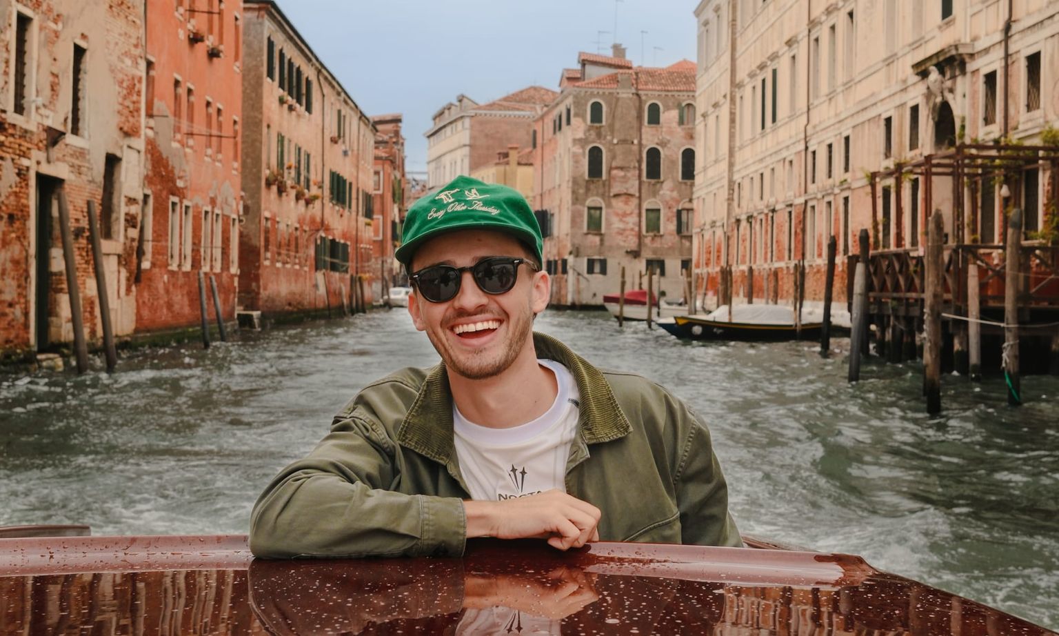 Smiling man in sunglasses and green cap on a boat, surrounded by Venice canals and historic buildings.