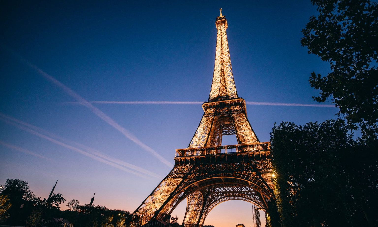 The Eiffel Tower illuminated at dusk, with a deep blue sky and light trails in the background, surrounded by silhouetted trees.