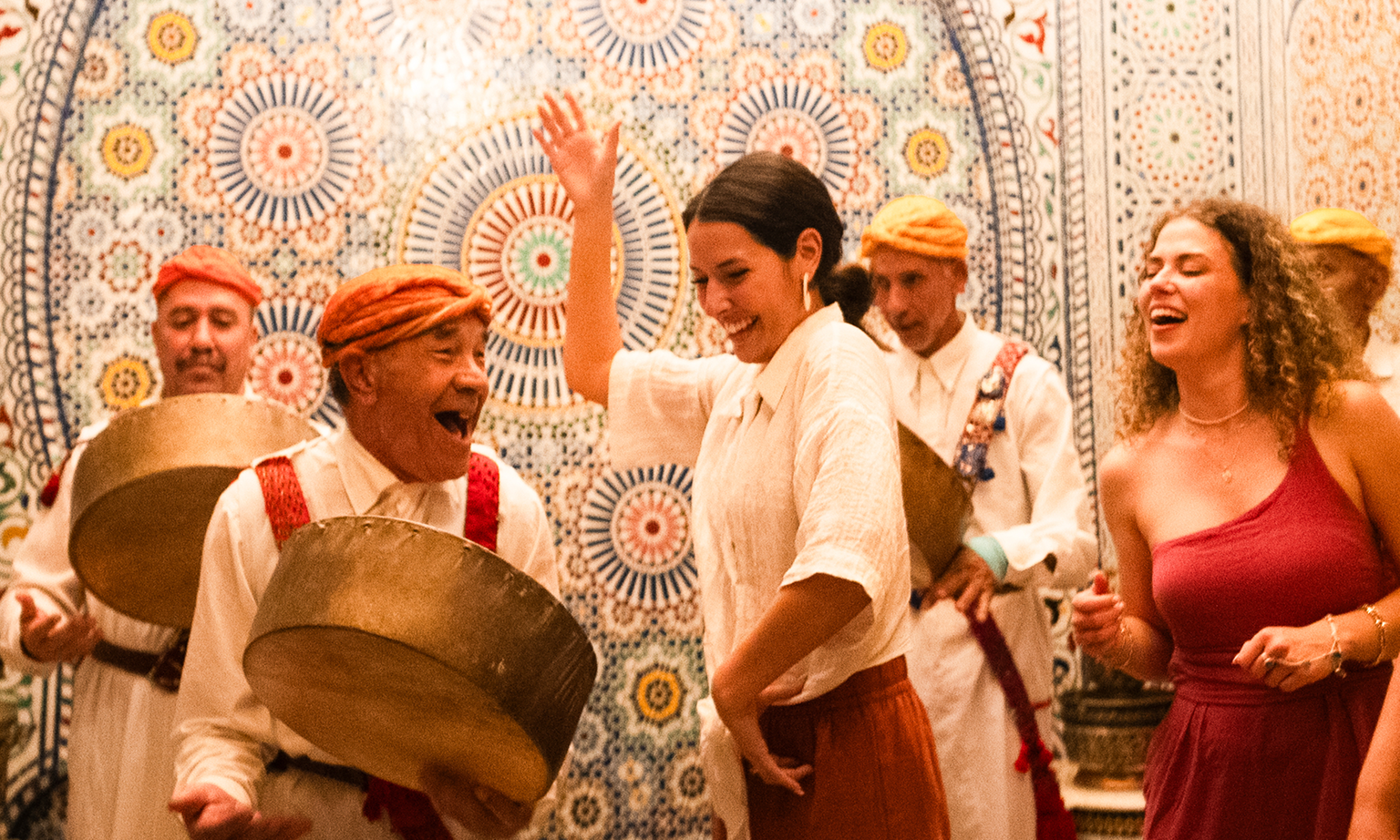 Four men banging handheld drums while two women dance in Morocco.