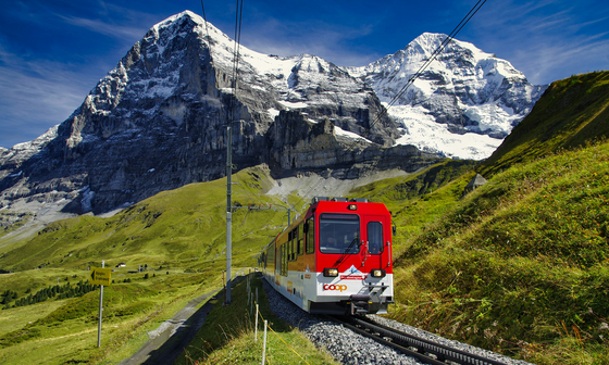 A red train travels on tracks through a mountainous landscape, with snow-capped peaks in the background and green grass in the foreground.