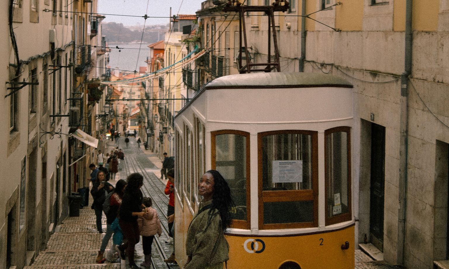 A tram ascending a narrow, hilly street lined with colorful buildings in Lisbon, with people walking nearby on a cloudy day.