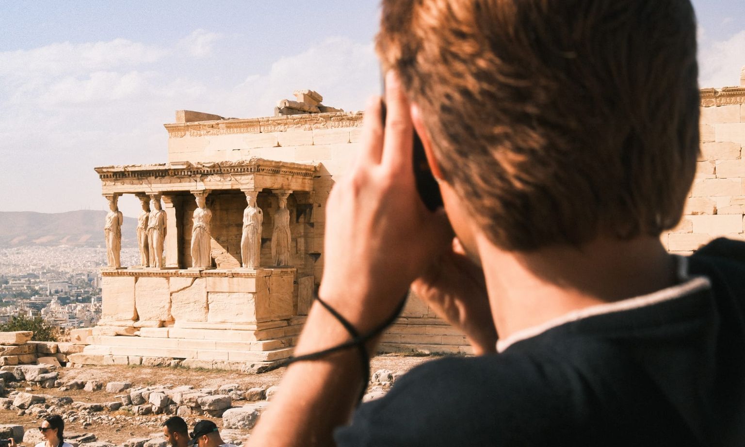 Person taking a photo of the Erechtheion with its Caryatids on the Acropolis in Athens, Greece, under a clear blue sky.