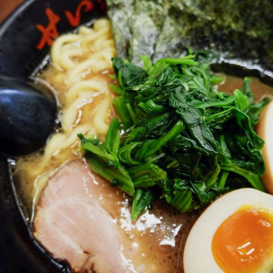 A bowl of ramen with sliced pork, soft-boiled eggs, seaweed, and greens in a rich broth, served with a black spoon.