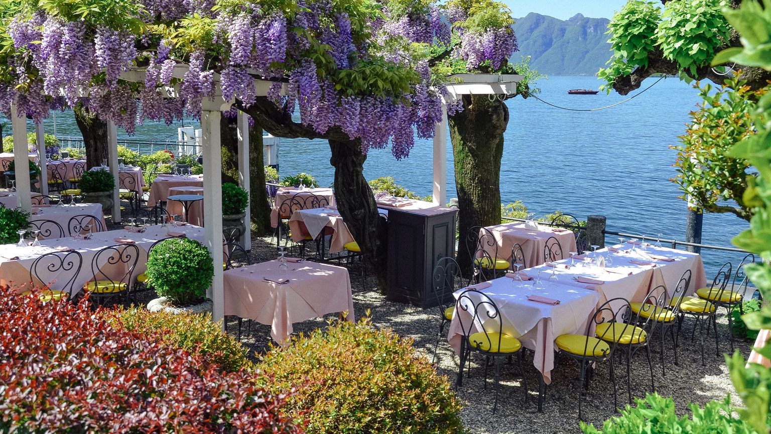 A restaurant with white tablecloths under a trellis of purple wisteria overlooking Lake Como, Italy