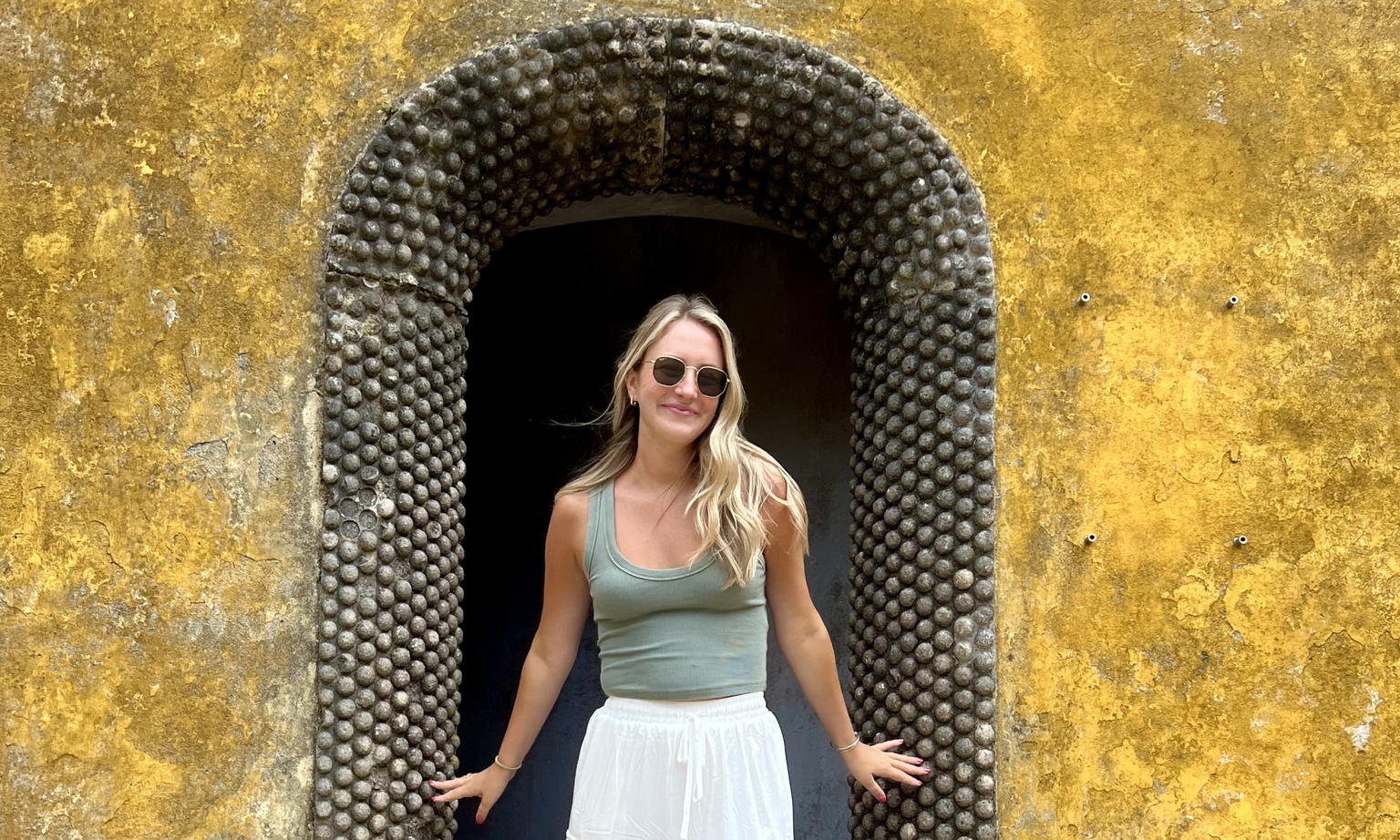 A woman in sunglasses smiling and posing under an arched stone door set in a bright yellow wall