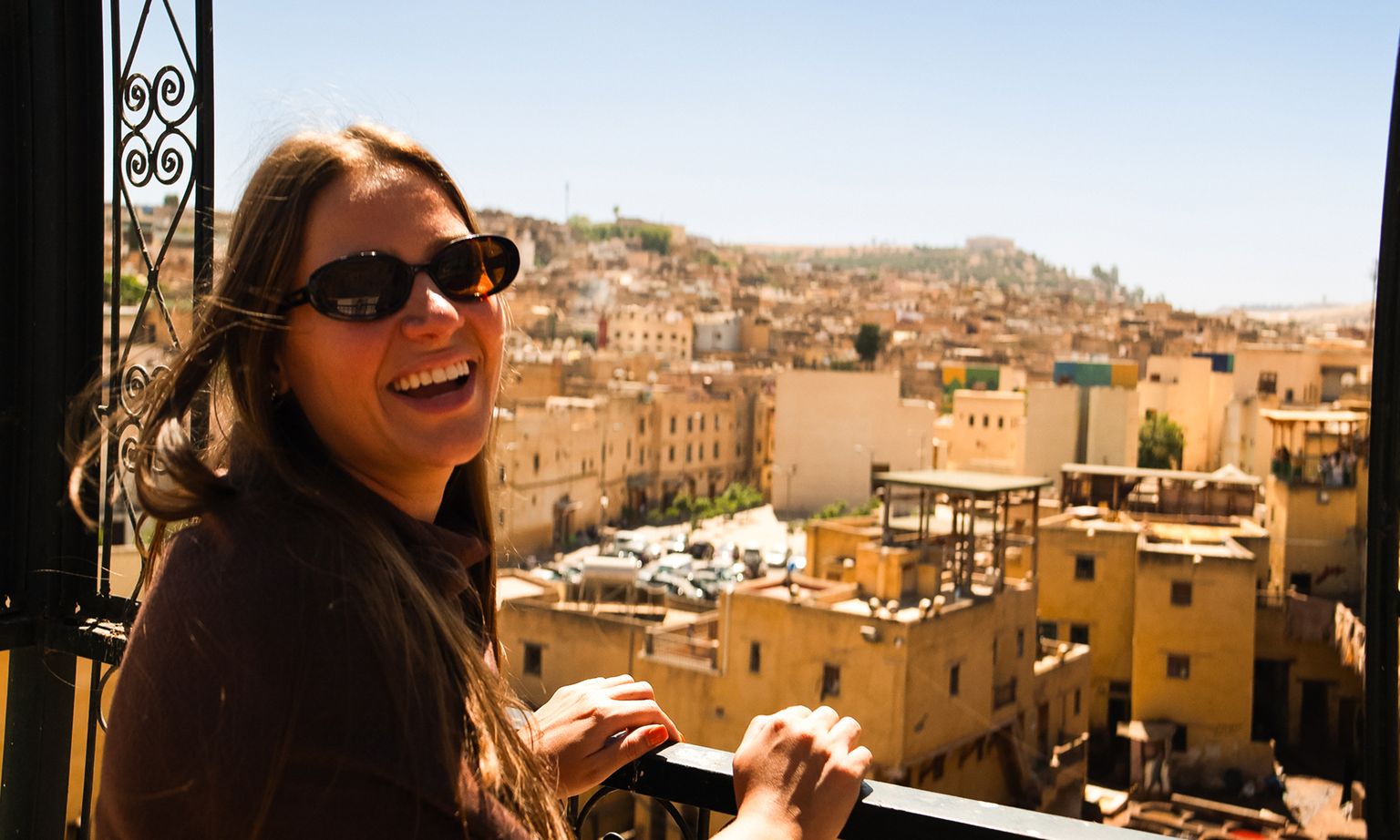 Smiling woman wearing sunglasses leans on a railing, overlooking a sunlit city with beige buildings and a clear blue sky.
