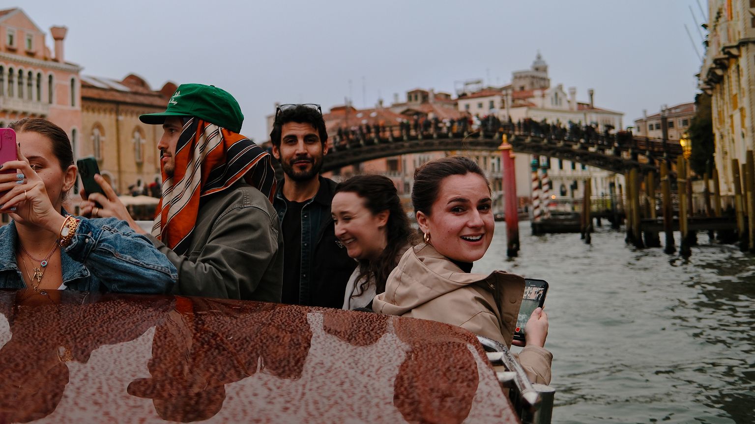 A group of people on a boat in Venice, capturing photos with their phones, with a canal and bridge in the background.
