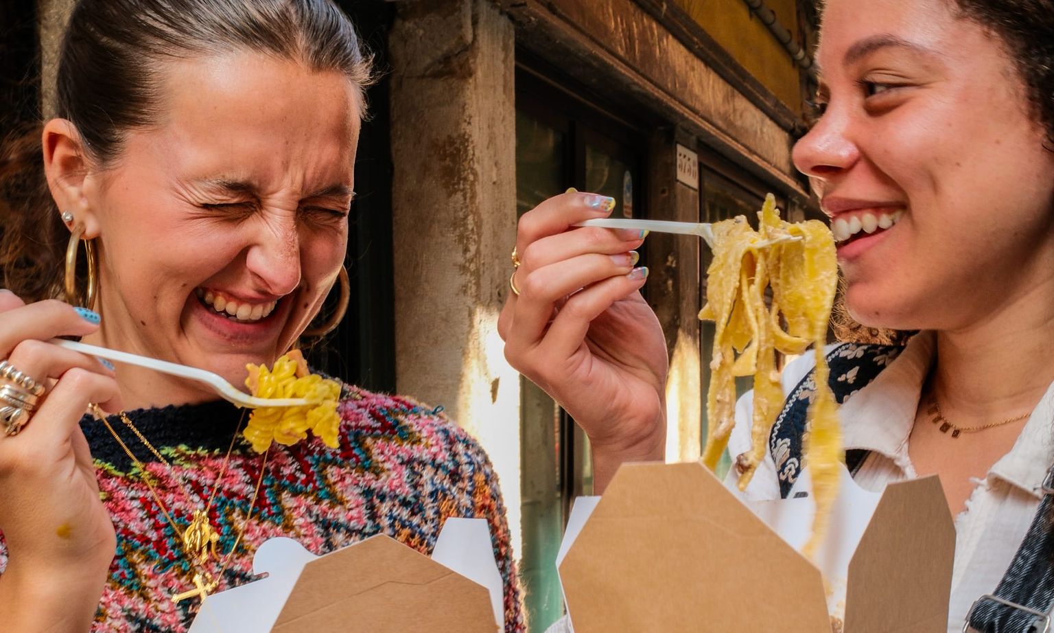Two people laughing and eating noodles from takeout boxes with plastic forks, standing outdoors and enjoying the moment.