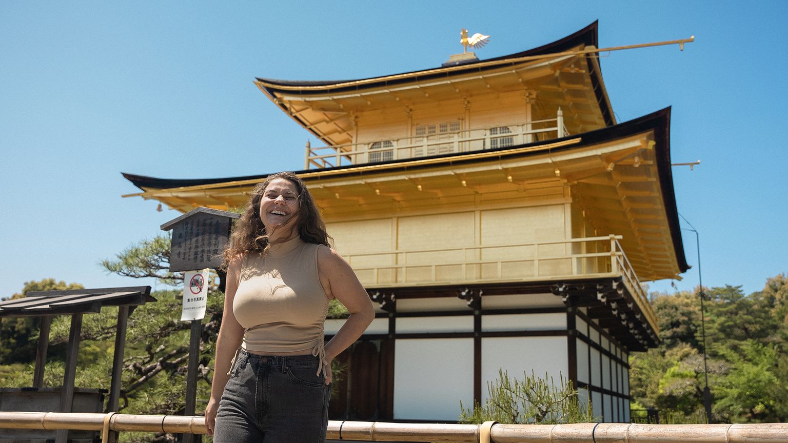 A woman smiles while standing in front of Kinkaku-ji, the Golden Pavilion in Kyoto, on a sunny day.