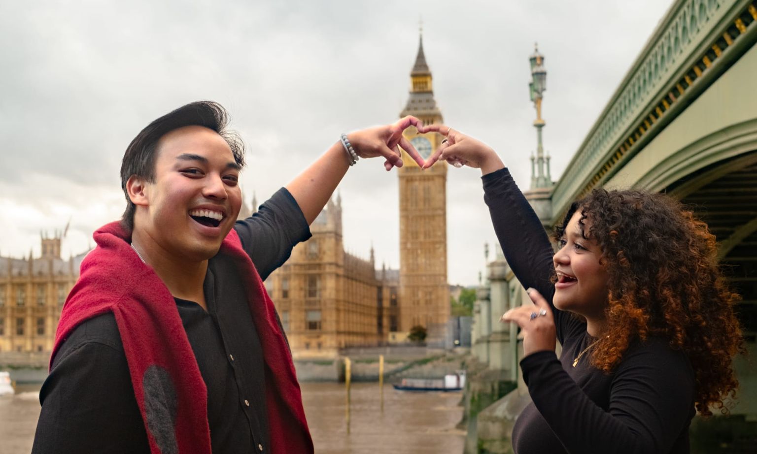 Two people smiling, forming a heart shape with their hands, with Big Ben and a bridge in the background. Overcast sky.