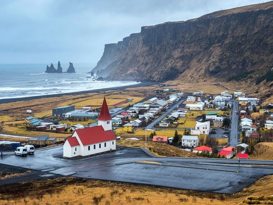 A small coastal village with a white church and red roof, surrounded by cliffs and ocean, under a cloudy sky.