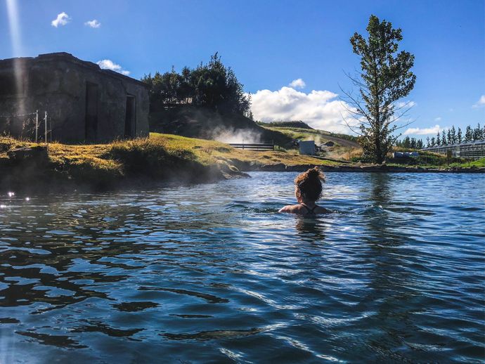 A person swims in a tranquil outdoor hot spring, surrounded by lush greenery, under a clear blue sky with fluffy clouds.