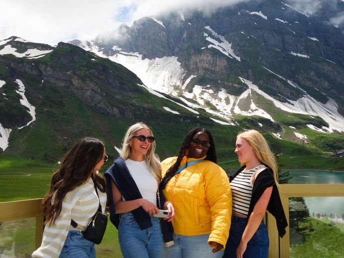 Four women smiling and laughing on a balcony with a scenic mountain backdrop, featuring snow-capped peaks and a lush green landscape.