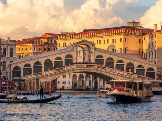 A view of the Rialto Bridge in Venice with boats and gondolas on the canal, set against a backdrop of historic buildings and a cloudy sky.