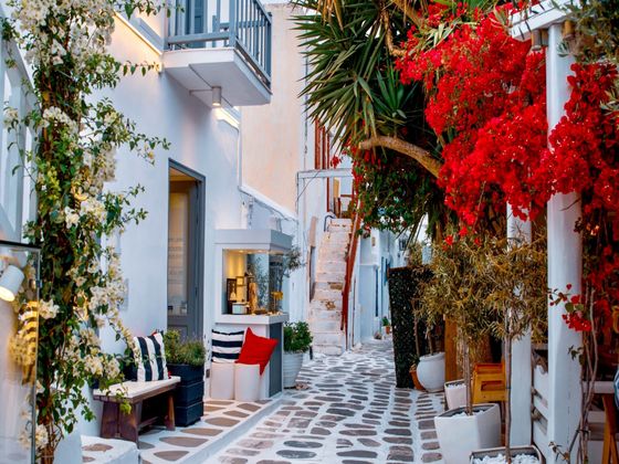 A charming narrow alley in a Greek village with whitewashed walls, flowering bougainvillea, potted plants, and cozy seating under a blue sky.