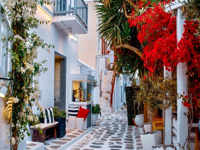 A charming narrow alley in a Greek village with whitewashed walls, flowering bougainvillea, potted plants, and cozy seating under a blue sky.