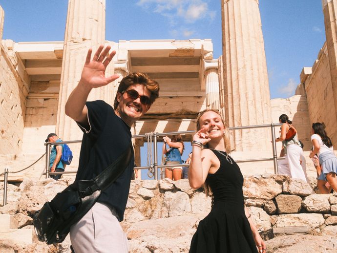A man and woman smiling and waving, standing in front of an ancient stone temple, with tourists in the background against a blue sky.