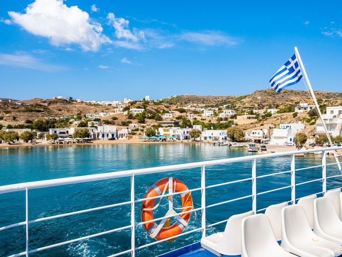 Ferry deck overlooking a Greek coastal village with white buildings, a Greek flag, and clear blue water under a sunny sky.
