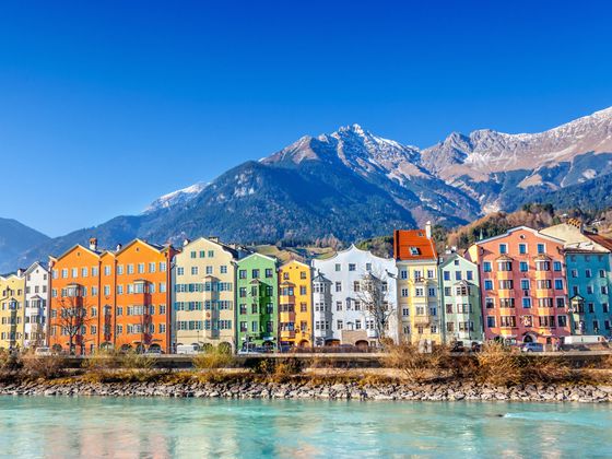 Colorful buildings line a riverbank with snow-capped mountains and a clear blue sky in the background.