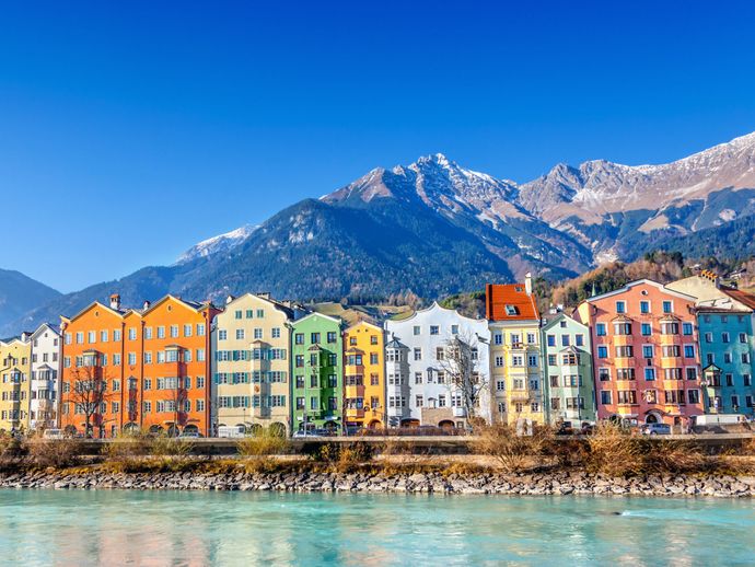 Colorful buildings line a riverbank with snow-capped mountains and a clear blue sky in the background.