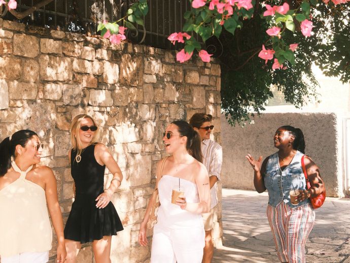 Five people smiling and chatting outdoors near a stone wall, with pink flowers hanging above, enjoying a sunny day.