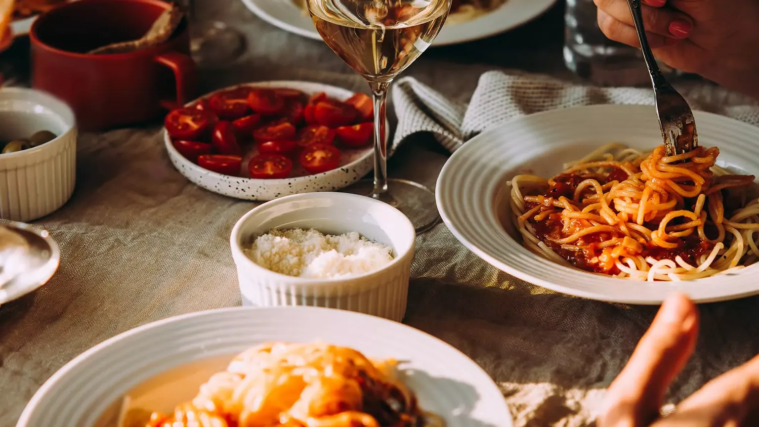 Table set with plates of spaghetti, glasses of white wine, a bowl of cherry tomatoes, and a dish of grated cheese. Hands holding forks.