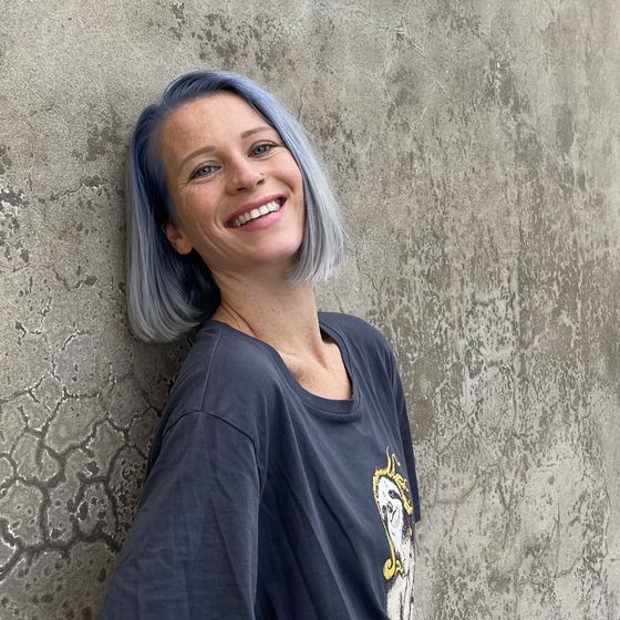 Smiling person with short blue-gray hair leans against a textured gray wall, wearing a dark shirt with a graphic print.
