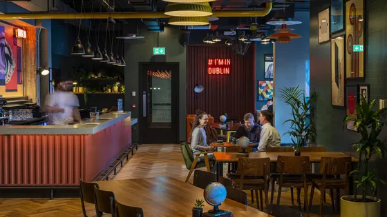 Contemporary cafe with colorful decor, neon sign "# I'm in Dublin," and three people seated at a table. Counter on the left with a blurred barista.