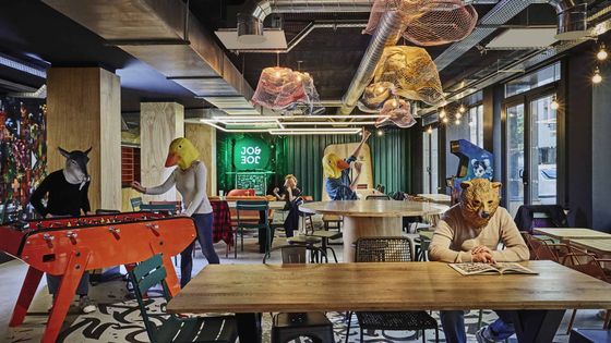 People in animal masks engage in activities like foosball and reading in a modern cafe with colorful decor and ample seating.