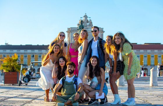 A group of smiling people posing together outdoors in a sunny plaza with a historical building in the background.