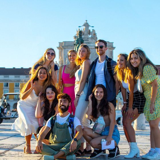 A group of smiling people posing together outdoors in a sunny plaza with a historical building in the background.