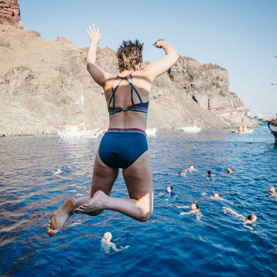 Person jumping into the sea from a boat, with swimmers nearby and a sailboat in the background, under a clear sky.