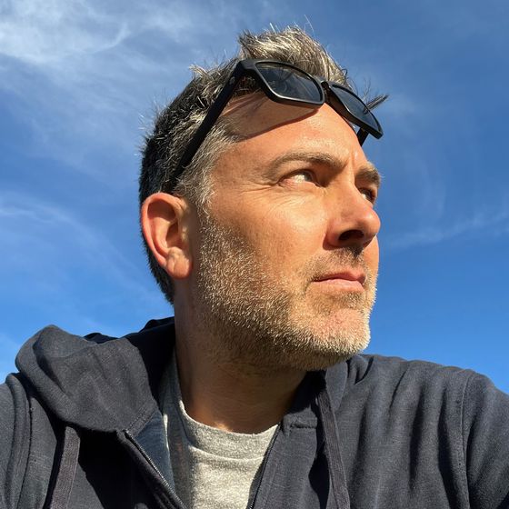 A man with short hair and sunglasses on his head looks into the distance against a clear blue sky, wearing a dark hoodie and a gray T-shirt.