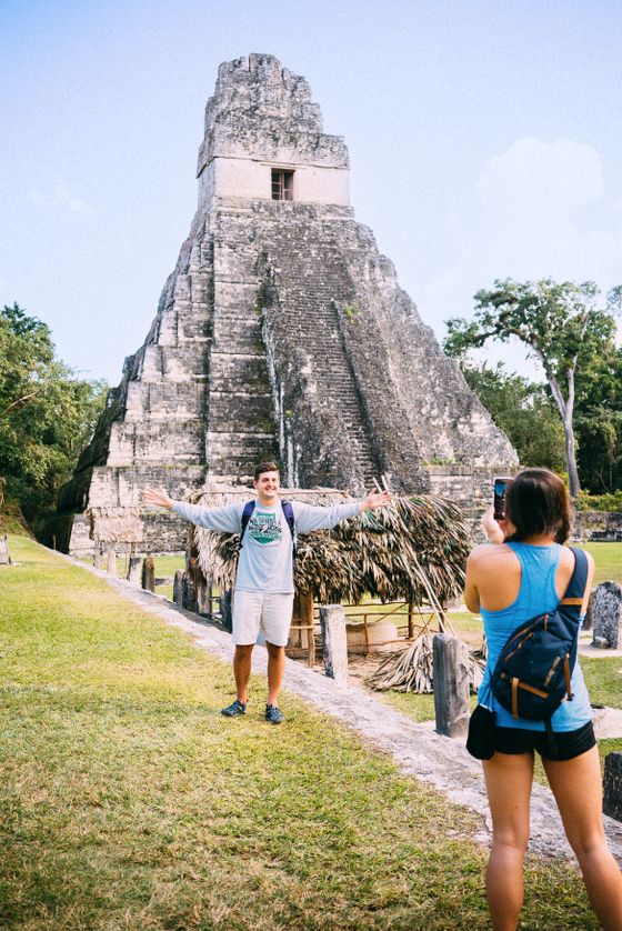 A person poses for a photo in front of a stone pyramid at Tikal, while another takes a picture, surrounded by lush greenery.