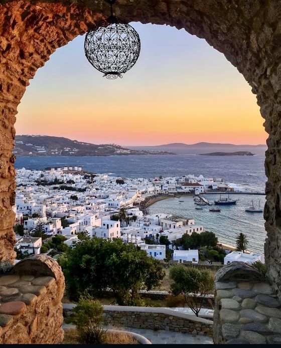 View through stone archway of a coastal town with white buildings at sunset, overlooking a tranquil bay with boats and distant hills.