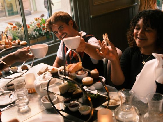People enjoying afternoon tea with a tiered tray of pastries and teapots on a table, smiling and raising their teacups in a cozy café setting.