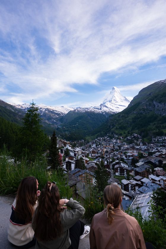 Three people sit on a grassy hill overlooking a small town with the snow-capped Matterhorn mountain in the background under a partly cloudy sky.
