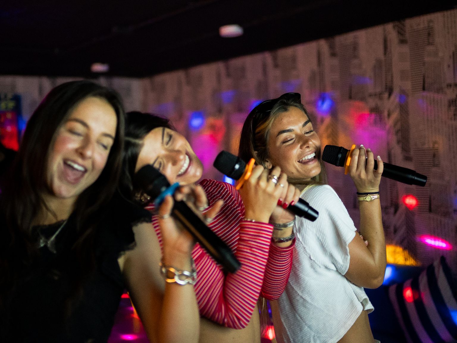 Three women joyfully sing into microphones in a dimly lit room with colorful lights, smiling and leaning close together.