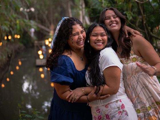 Three women smiling and embracing in a lush, wooded area by a waterway with warm lights in the background.