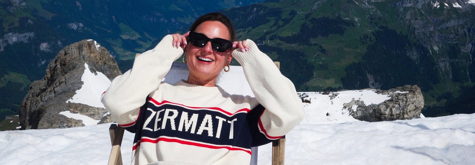 A person wearing sunglasses and a "Zermatt" sweater sits on a snowy mountain, smiling, with a scenic view of mountains in the background.