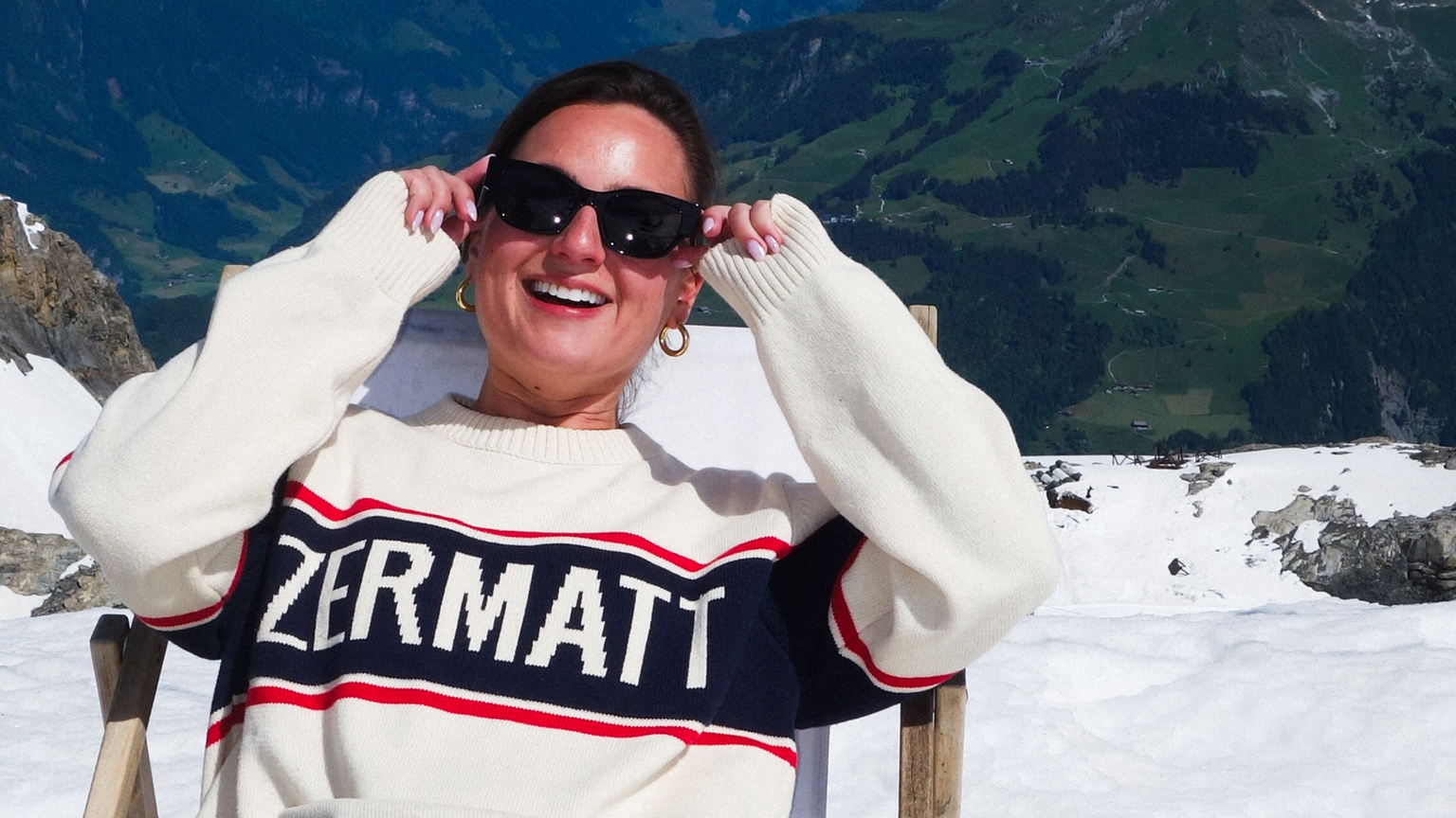 A person wearing sunglasses and a "Zermatt" sweater sits on a snowy mountain, smiling, with a scenic view of mountains in the background.