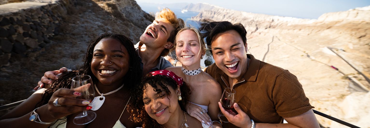 A group of five friends, smiling and holding wine glasses, pose for a selfie in a sunny, rocky outdoor setting with mountains in the background.