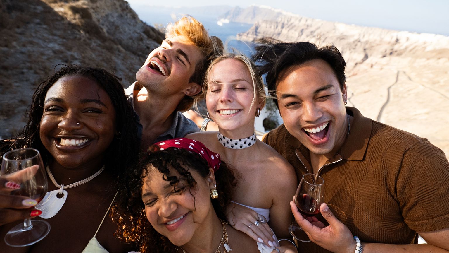 A group of five friends, smiling and holding wine glasses, pose for a selfie in a sunny, rocky outdoor setting with mountains in the background.