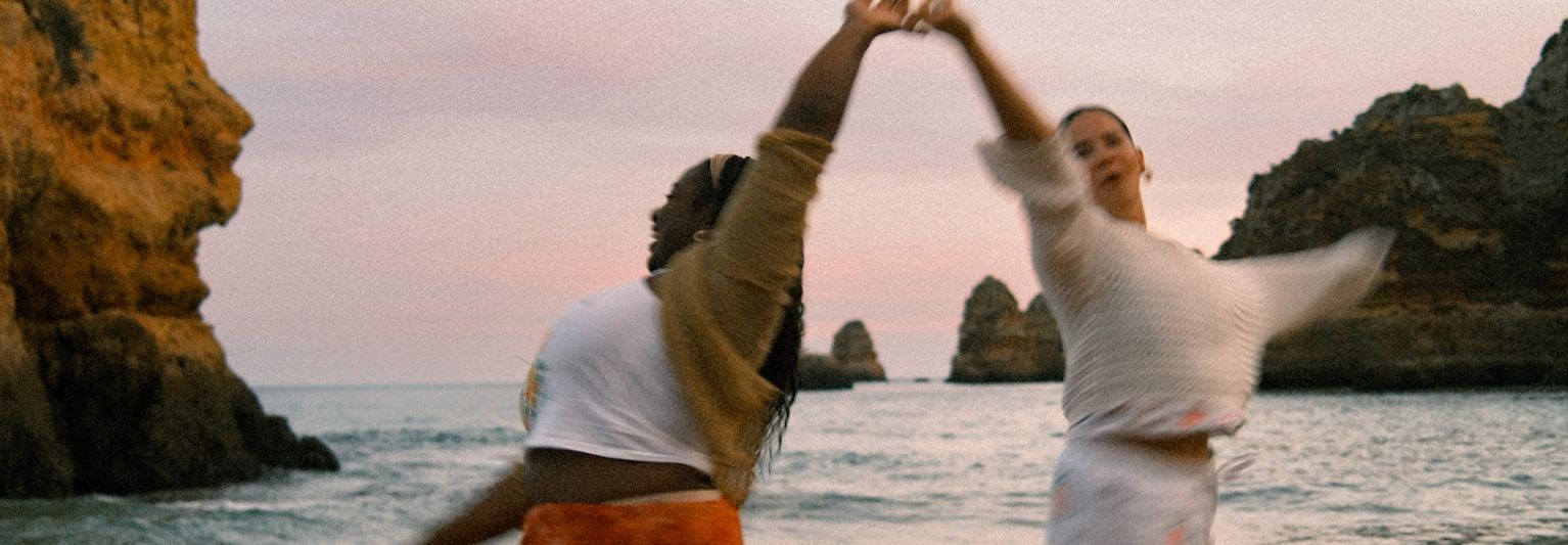 Two people joyfully dancing by the sea at sunset, surrounded by rocky cliffs and gentle waves in the background.